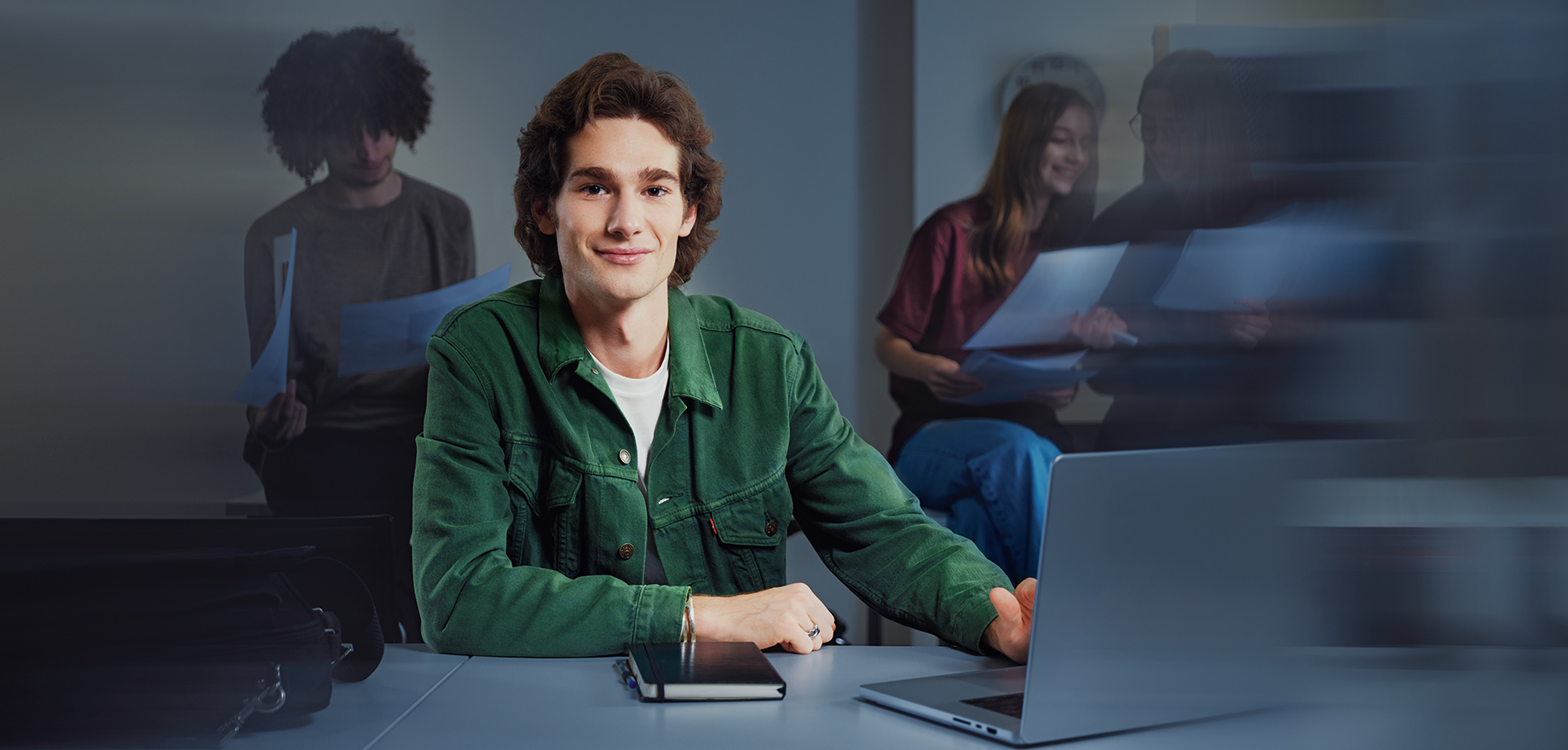 Junger Mann mit einem Laptop sitzt am Tisch und l&auml;chelt.