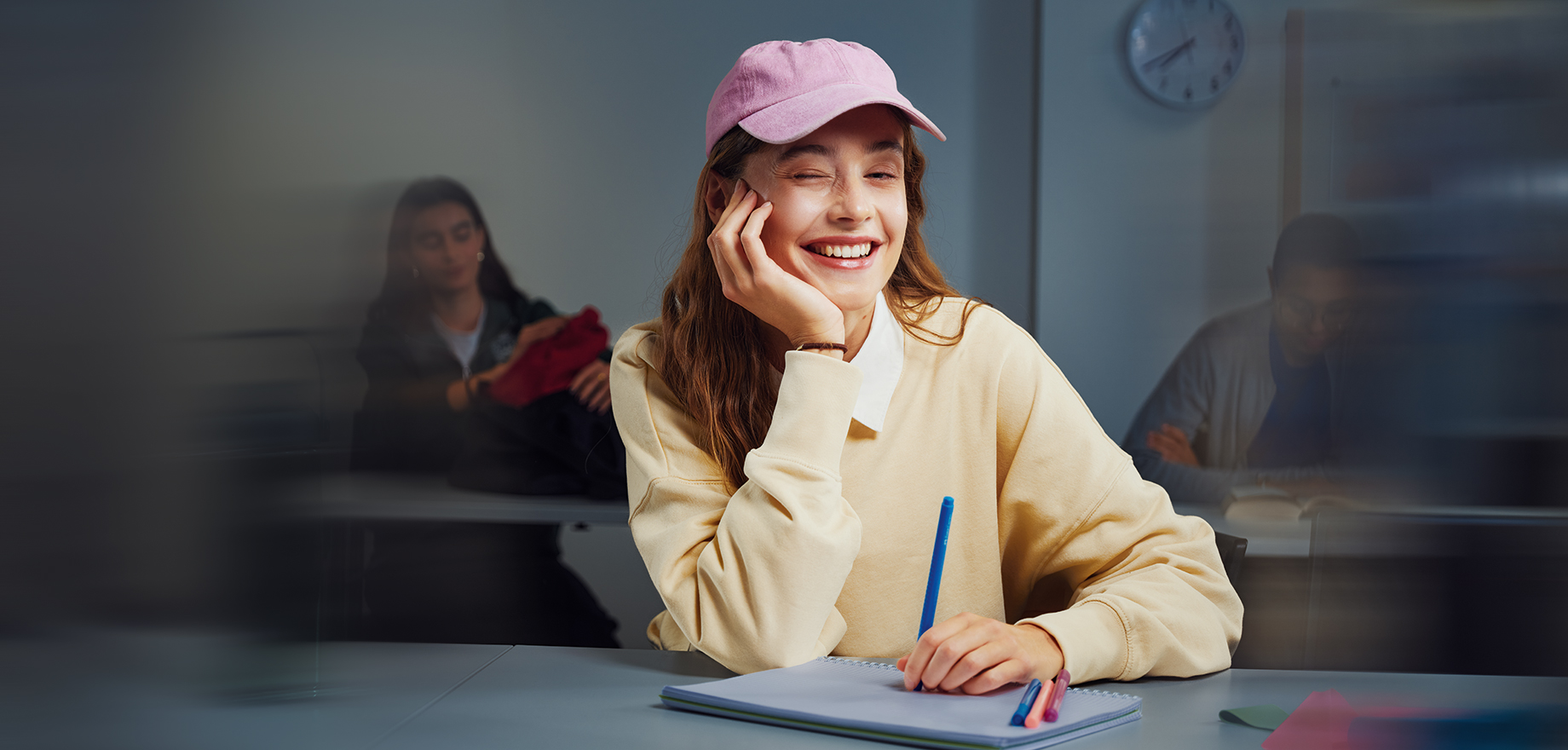 Junge Frau mit Cap l&auml;chelt in die Kamera und sitzt an einem Tisch mit Block und Stift